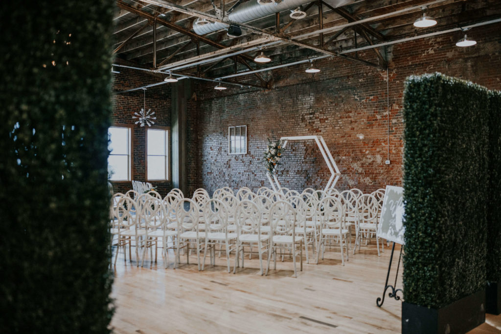 Rustic industrial indoor wedding ceremony space with white geometric wedding arch and chairs 
