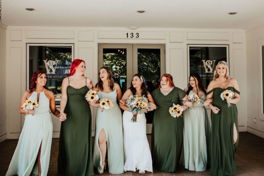 Bride and bridesmaids wearing shades of green dresses and carrying sunflower bouquets outside of White Star Station wedding venue